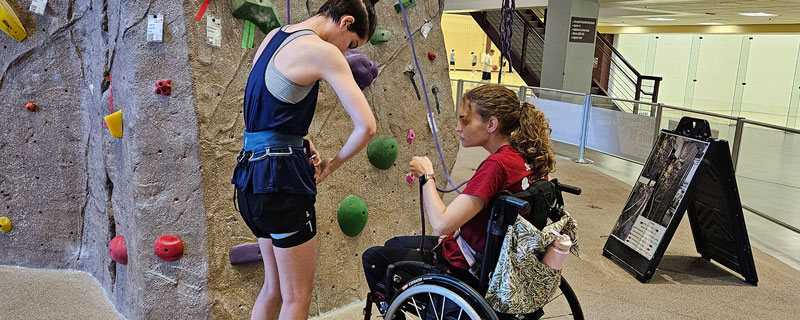 Adaptive climber, sitting in her wheelchair learning to tie in to the climbing rope while at the base of the rockwall with a climbing instructor.