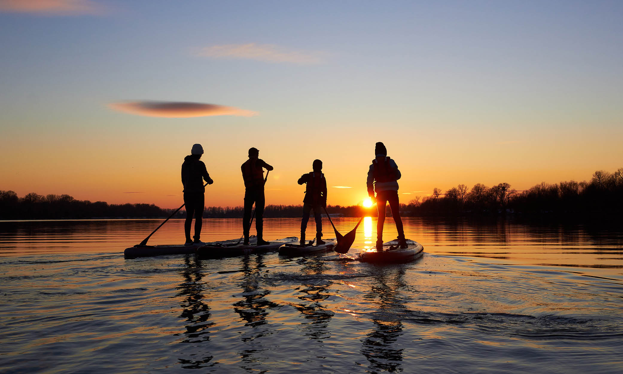 Four people are in a lake standing on a paddle board with a sunset in the background.