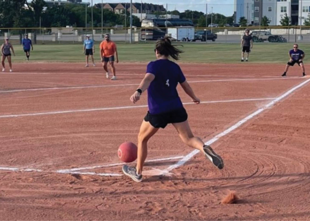Sports_kickball A person in a blue shirt and black shorts is kicking a red soccer ball on a dusty field, with several other players visible in the background.