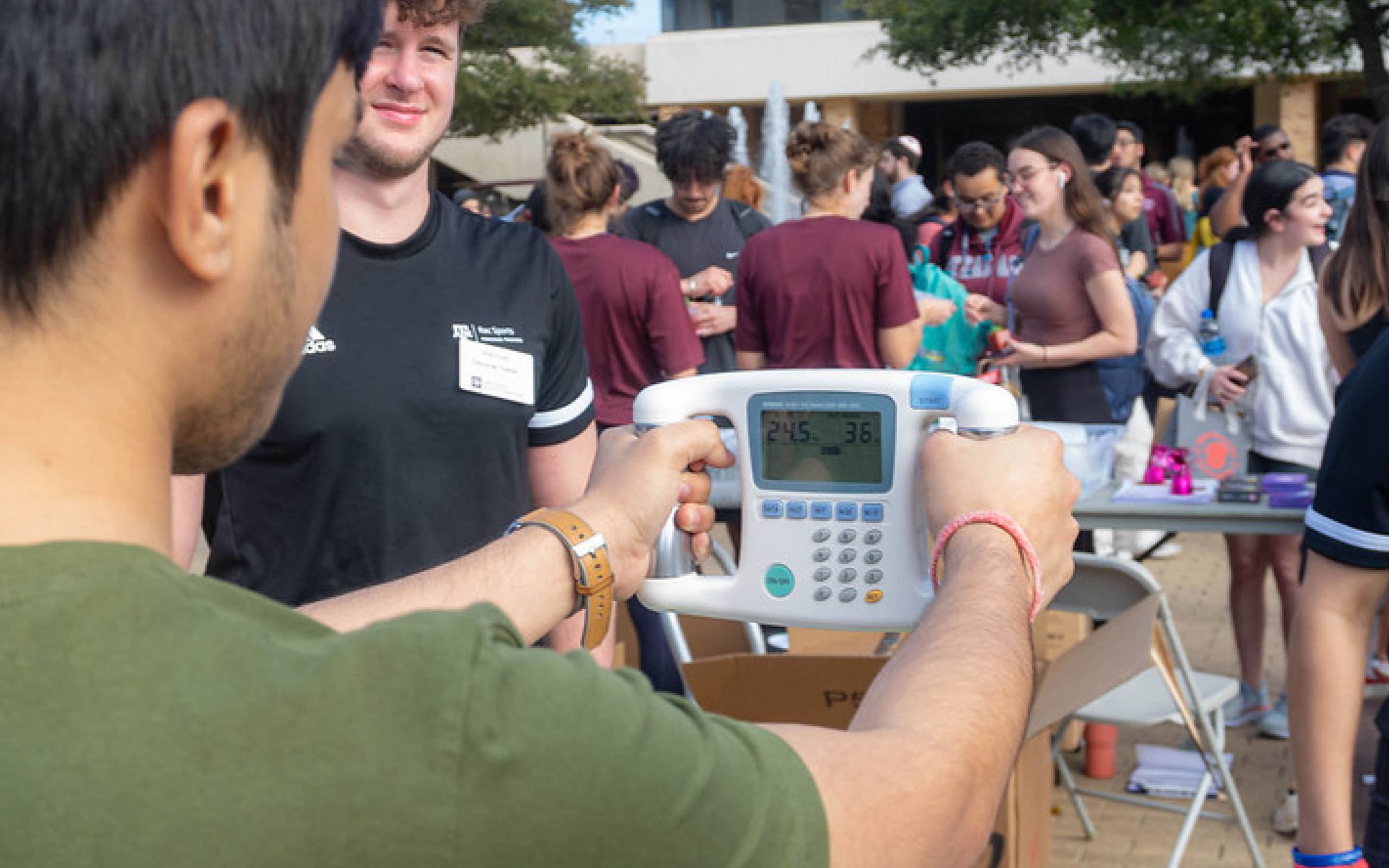 An individual holding a calculator displaying the number 26.36 at a busy outdoor event with people conversing in the background.