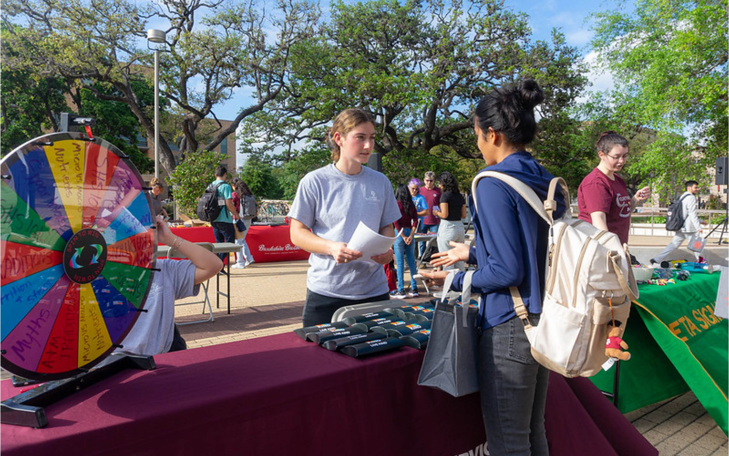 Two people are engaging in a conversation at a university promotional booth, where various giveaway items are displayed on the table. In the background, there is a colorful prize wheel and additional participants at the event.