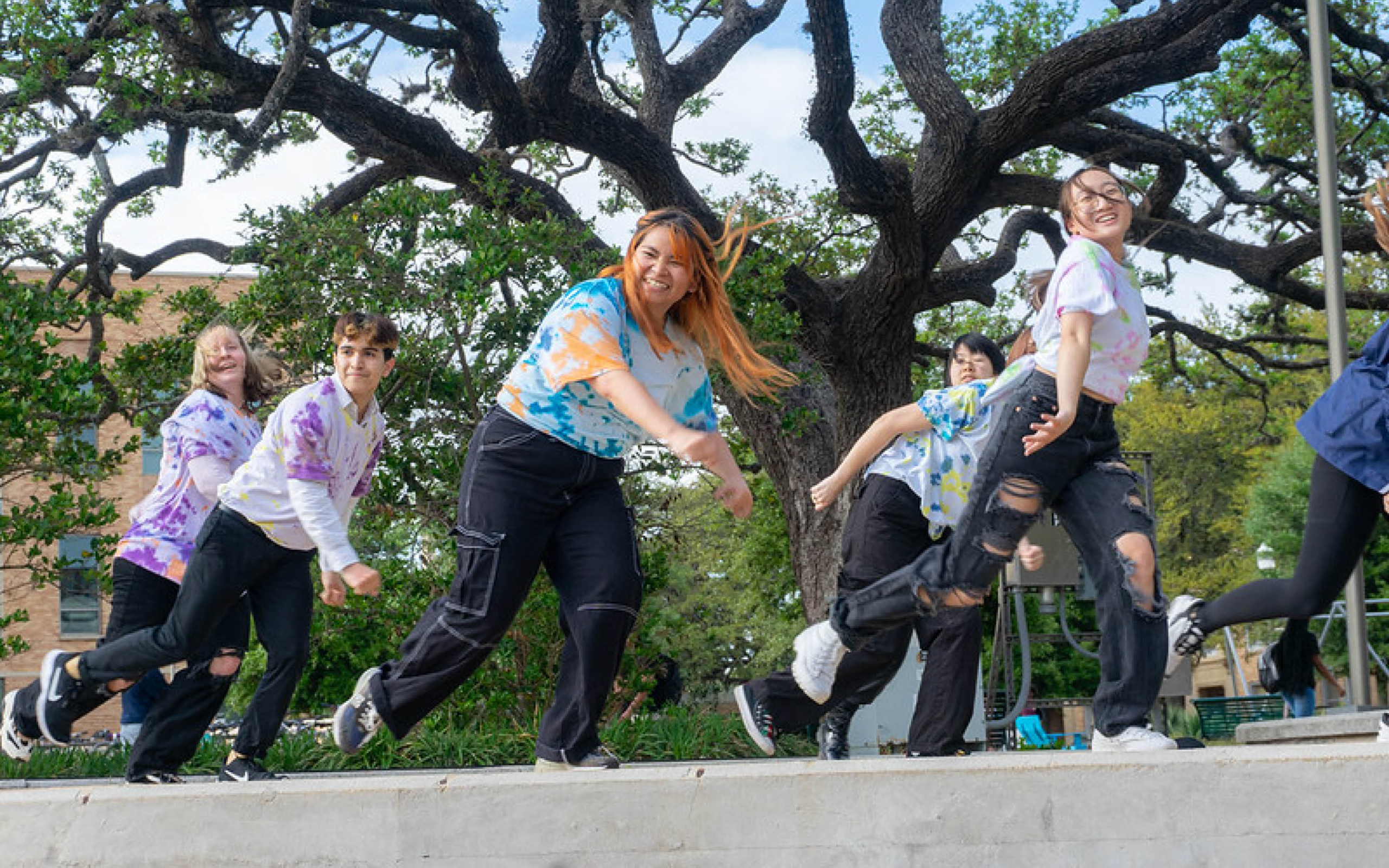 A group of individuals, wearing colorful tie-dye shirts, joyfully leaping and smiling in front of a large oak tree.