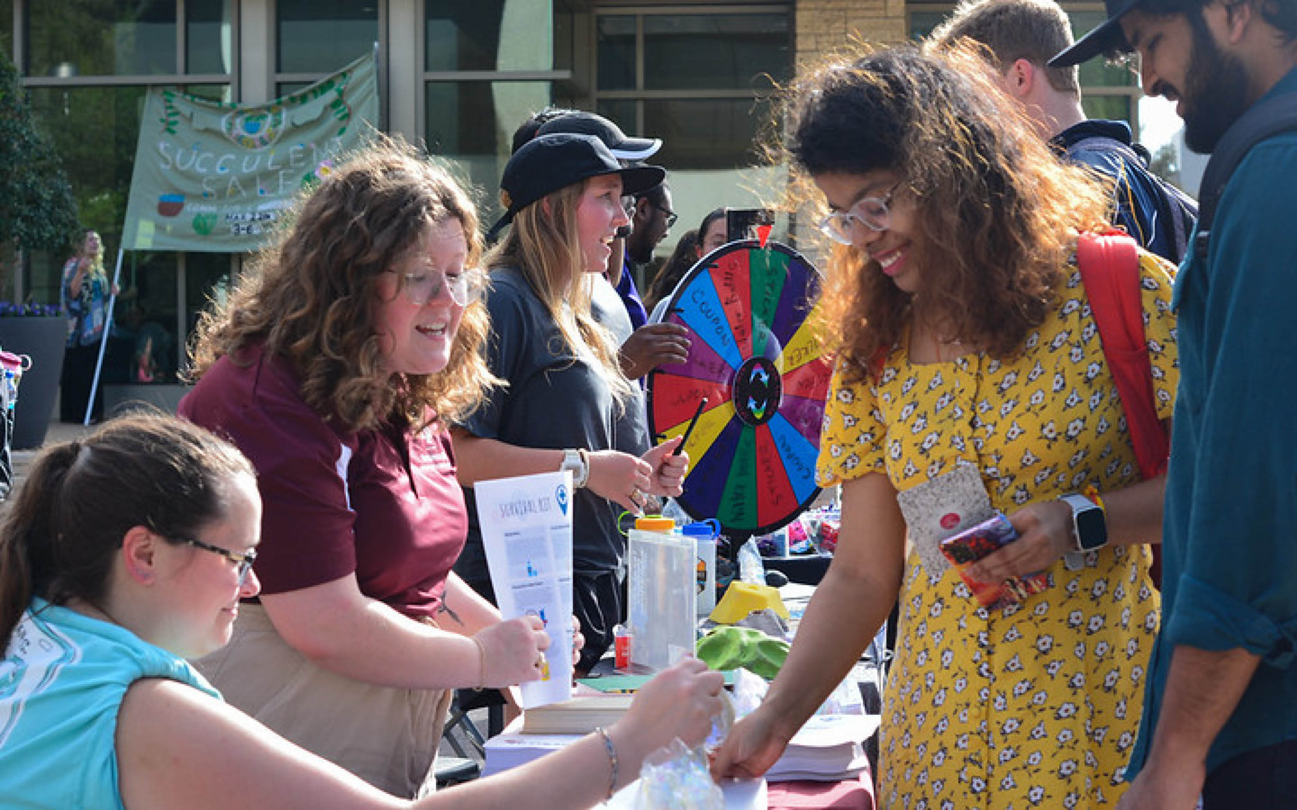 Attendees interacting at a university promotional event with a prize wheel and informational booths in the background.