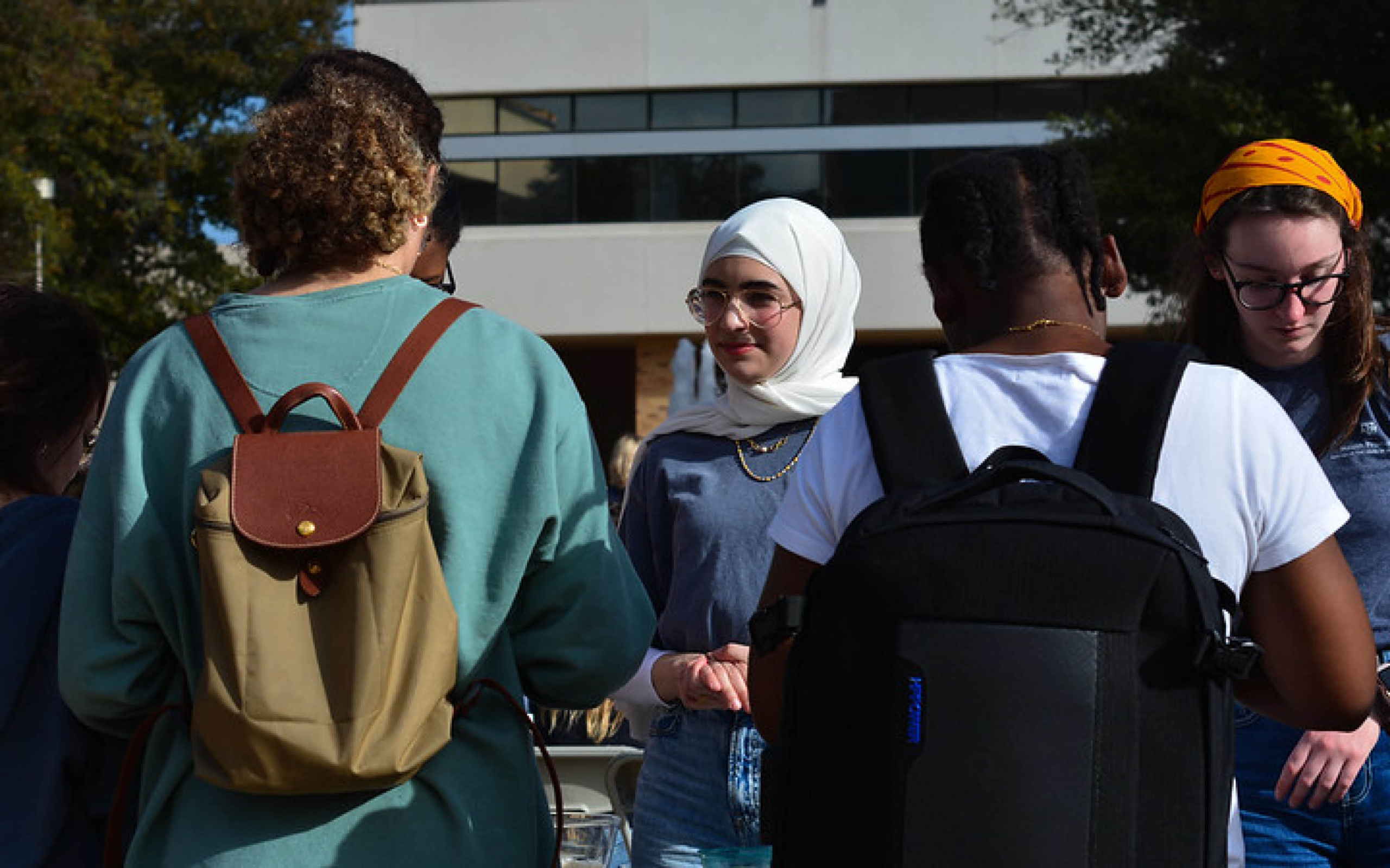 Group of students conversing outdoors, one wearing a hijab, with backpacks visible.